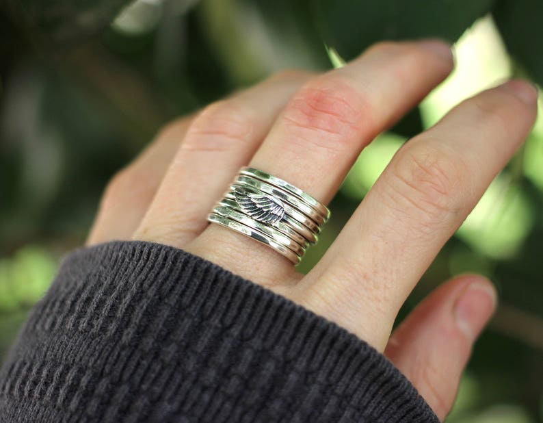 May include: A silver ring with a stack of bands featuring a small wing design. The ring is worn on a person's finger.