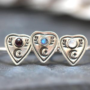 May include: Three silver rings with heart-shaped charms. Each charm features a crescent moon and a star, and is adorned with a different gemstone: a red garnet, a blue moonstone, and a white moonstone.