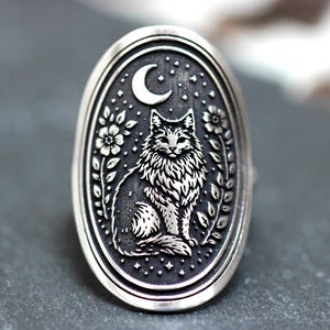 Op de afbeelding: Zilverkleurige ring met een ovale voorkant met een gedetailleerd kattenontwerp. De kat zit onder een maan en sterren, omringd door bloemmotieven. De ring heeft een getextureerde, antieke afwerking.