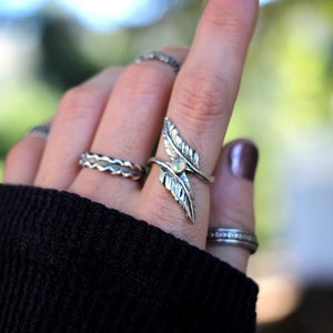 May include: A silver ring with a leaf design and a small opal stone. The ring is worn on the index finger of a hand.