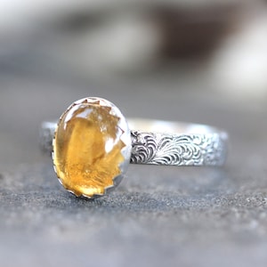 May include: A silver ring with a yellow gemstone set in a scalloped bezel. The band is decorated with a floral pattern.