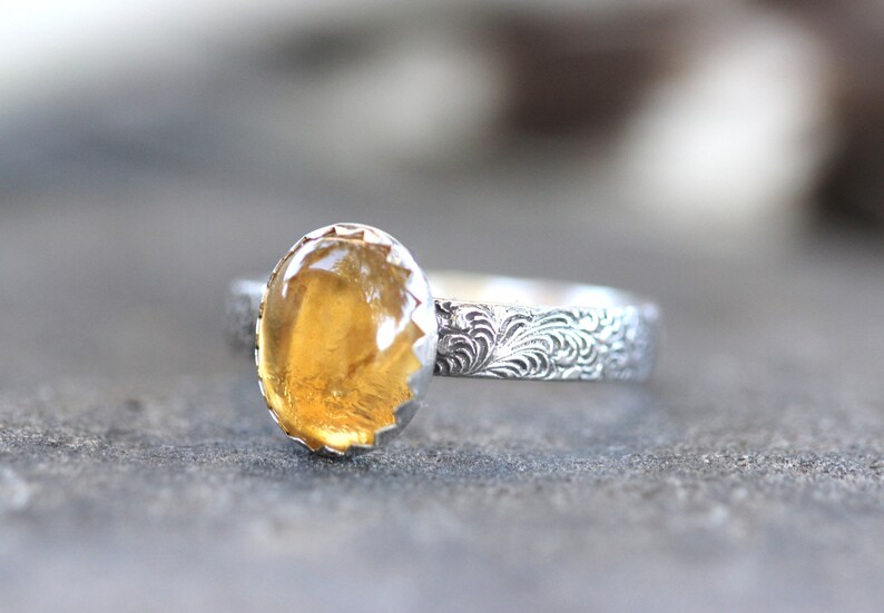 May include: A silver ring with a yellow gemstone set in a scalloped bezel. The band is decorated with a floral pattern.
