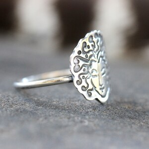 May include: A silver ring with an oval face featuring a sun design. The ring's face has a detailed pattern around the sun's face. The ring is on a grey surface.