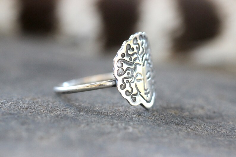 May include: A silver ring with an oval face featuring a sun design. The ring's face has a detailed pattern around the sun's face. The ring is on a grey surface.