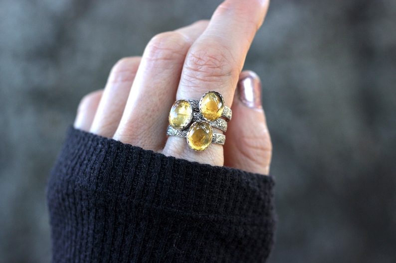 May include: Three silver rings with yellow oval gemstones. The rings are stacked on a finger.