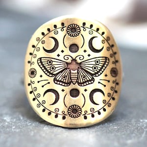 May include: A gold-colored ring with a detailed moth and crescent moon design. The ring features an oval shape with intricate etched patterns of moons, flowers, and a central moth. The ring is likely made of metal.