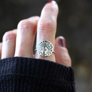 May include: Silver ring with a detailed face design and flowing hair. The ring is worn on a finger, highlighting the intricate details of the jewellery. The ring's design is centered on a round shape, with a face in the middle.