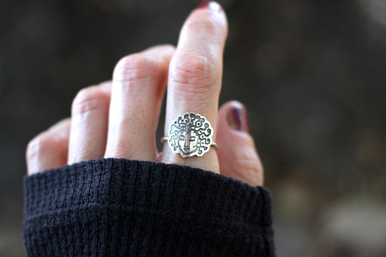 May include: Silver ring with a detailed face design and flowing hair. The ring is worn on a finger, highlighting the intricate details of the jewellery. The ring's design is centered on a round shape, with a face in the middle.
