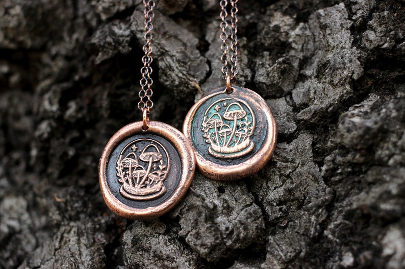 May include: Two copper necklaces with mushroom pendants. The pendants are round and have a green enamel background with a design of mushrooms and leaves. The chains are silver.
