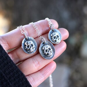 May include: Three silver skull pendant necklaces. Each pendant features a detailed skull within an oval frame, suspended from a delicate chain. The necklaces are held in a hand, showcasing the jewelry's design and craftsmanship.