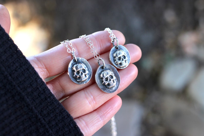 May include: Three silver skull pendant necklaces. Each pendant features a detailed skull within an oval frame, suspended from a delicate chain. The necklaces are held in a hand, showcasing the jewelry's design and craftsmanship.