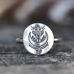 May include: A silver ring with a round face featuring an etched design of a crescent moon, a crystal, and a stylized arrow.