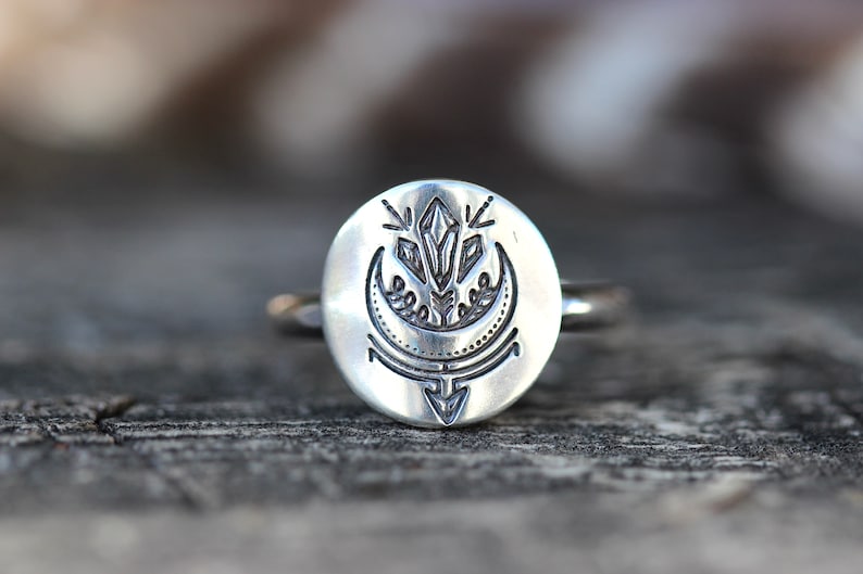 May include: A silver ring with a round face featuring an etched design of a crescent moon, a crystal, and a stylized arrow.