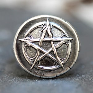 Puede incluir: Anillo plateado con un diseño de pentagrama en relieve dentro de un marco circular. Las puntas de la estrella son alargadas y texturizadas, sobre un fondo envejecido y detallado. El anillo tiene un aspecto desgastado.
