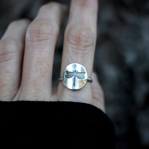 May include: A silver ring featuring a circular face with a detailed dragonfly design. The dragonfly is centered on the ring's face, with its wings spread. The ring is worn on a finger, with a blurred background.