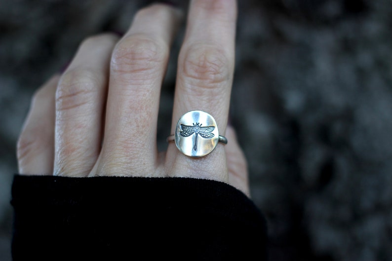 May include: A silver ring featuring a circular face with a detailed dragonfly design. The dragonfly is centered on the ring's face, with its wings spread. The ring is worn on a finger, with a blurred background.