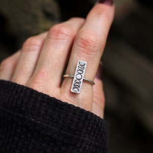 May include: A silver ring with a vertical design of eight moon phases. The ring is worn on a finger.