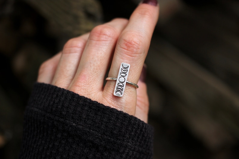 May include: A silver ring with a vertical design of eight moon phases. The ring is worn on a finger.
