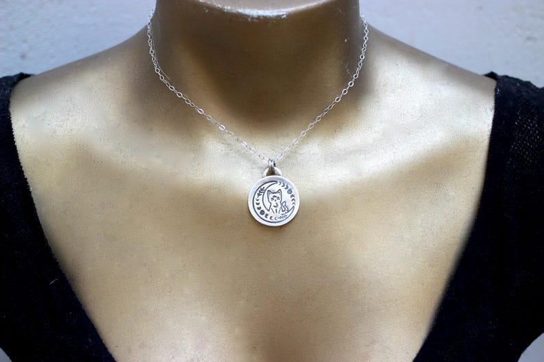 May include: A silver necklace with a pendant featuring a cat sitting on a crescent moon. The pendant is detailed with stars and other celestial symbols.