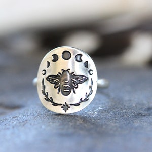 May include: A silver ring with an oval-shaped design featuring a bee and a crescent moon with stars.