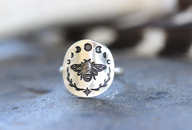 May include: A silver ring with an oval-shaped design featuring a bee and a crescent moon with stars.