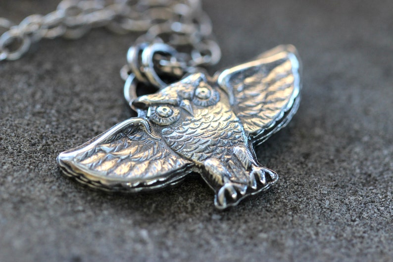 May include: Silver owl pendant with detailed wings and eyes. The owl is perched with its wings spread out.