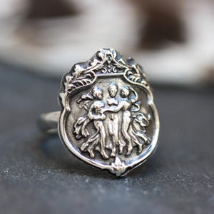 Puede incluir: Un anillo de plata con un dise&ntilde;o detallado que presenta tres figuras tomadas de la mano. Las figuras est&aacute;n rodeadas por un borde decorativo con motivos florales.