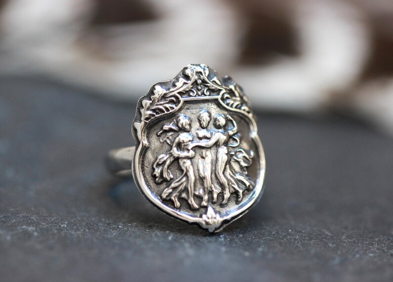 Puede incluir: Un anillo de plata con un dise&ntilde;o detallado que presenta tres figuras tomadas de la mano. Las figuras est&aacute;n rodeadas por un borde decorativo con motivos florales.