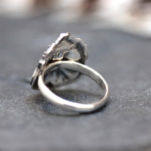Puede incluir: Un anillo de plata con un dise&ntilde;o detallado en la parte superior. El anillo est&aacute; sobre una superficie gris.