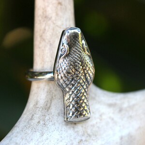 May include: A silver snake head ring with a detailed textured design. The ring is worn on a white surface.
