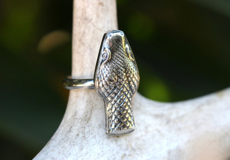 May include: A silver snake head ring with a detailed textured design. The ring is worn on a white surface.