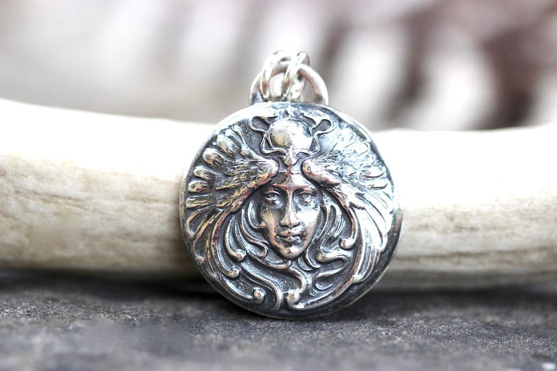 May include: A silver pendant featuring a detailed, stylised image of a woman's face with wings. The pendant is round and has a textured, ornate design.