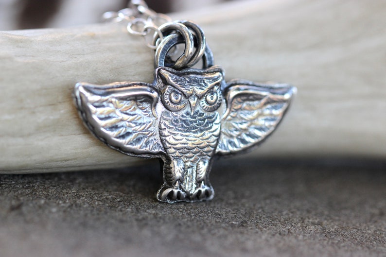 May include: A silver owl pendant with detailed wings and a textured surface. The owl has large, round eyes and a beak.