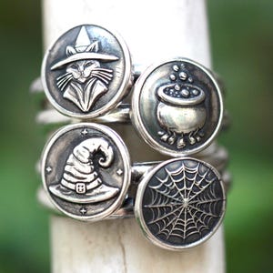 May include: A stack of silver rings with Halloween-themed designs. Each ring features a circular emblem with a different embossed image: a witch's cat, a bubbling cauldron, a witch's hat, and a spiderweb. The rings are displayed against a blurred green background.