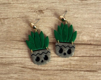 Puede incluir: Un par de pendientes hechos a mano con un diseño de suculenta. Cada pendiente tiene un perno de color dorado, una suculenta verde oscuro y una maceta gris con una cara de calavera. Los pendientes están colocados sobre un fondo de madera.