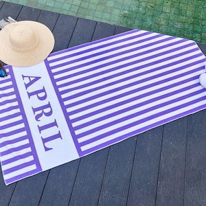 May include: A purple and white striped beach towel with the word "APRIL" printed in large letters. A straw sun hat, sunglasses, and white slippers are placed on the towel. The towel is on a wooden deck.