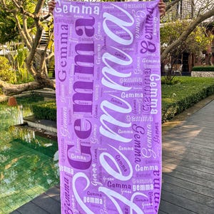 May include: A purple beach towel with the name "Gemma" printed repeatedly in white and purple. The towel is rectangular and held up against a background of a pool and trees.