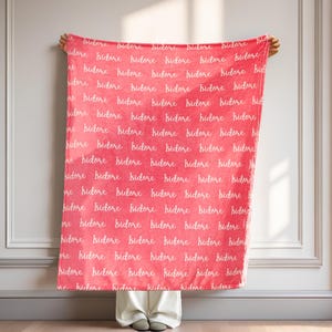 Op de afbeelding: Een koraalkleurige deken met het woord "bidore" in witte letters herhaald. De deken wordt getoond, waarbij het patroon en de kleur zichtbaar zijn. Een zacht, pluche woondecoratie-artikel.