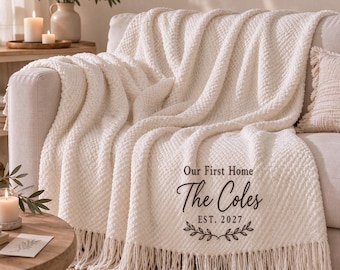 May include: A cream-colored textured throw blanket with fringe trim, featuring the printed text "Our First Home The Coles EST. 2027" accompanied by a small leaf graphic in dark brown ink, draped over a sofa.