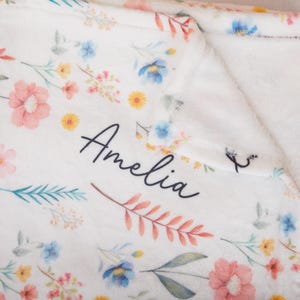 May include: A white blanket adorned with a floral pattern and the name "Amelia". The design includes pink, blue, yellow, and green flowers. The blanket has a soft, plush texture, perfect for comfort and warmth.