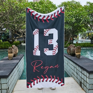 May include: A black baseball-themed blanket with the number 13 in white and the name "Regan" in red script. The blanket features red stitching details and a baseball design along the top and bottom edges. The blanket is displayed outdoors.