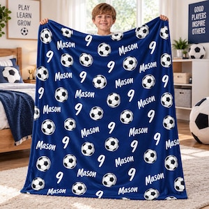 Op de afbeelding: Een donkerblauwe fleece deken met een herhalend patroon van zwart-witte voetballen, het cijfer 9 en de naam Mason in wit handschrift. De deken wordt omhoog gehouden om het volledige ontwerp over het gehele oppervlak te tonen.