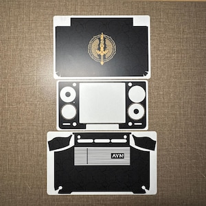 May include: A set of three black adhesive vinyl skins for a handheld gaming console. The top piece features a gold sword emblem. The middle piece has cutouts for a screen and buttons. The bottom piece includes horizontal vents and the text AYN.