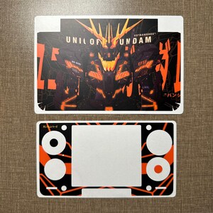 Peut inclure: Une feuille d'autocollants rectangulaire présentant un graphique violet foncé et orange du Unicorn Gundam 02 Banshee avec le texte UNICORN GUNDAM et 02 BANSHEE. En dessous se trouve une protection de console assortie avec découpes.