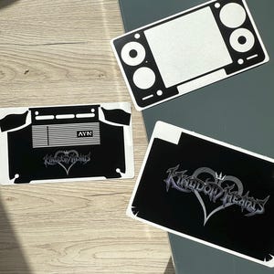 Könnte beinhalten: Drei schwarz-weiße Vinyl-Aufkleber für ein Handheld-Spielgerät. Ein Aufkleber hat einen Bildschirmausschnitt, ein weiterer zeigt das AYN-Logo mit horizontalen Linien und der dritte zeigt das Kingdom Hearts-Logo mit einem Herz- und Kronensymbol.