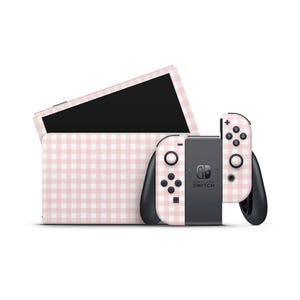 Puede incluir: Una consola Nintendo Switch y sus mandos con un patrón de cuadros rosa y blanco. La consola está en su base, y los mandos están conectados. El logotipo de Nintendo Switch es visible en la consola.
