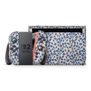 Könnte beinhalten: Eine Nintendo Switch-Konsole und Controller mit floralem Design. Das Design zeigt blaue Blumen und Blätter auf cremefarbenem Hintergrund. Die Controller haben schwarze Knöpfe und eine korallenfarbene Schiene.