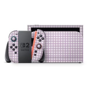 Könnte beinhalten: Eine Nintendo Switch-Konsole und Controller mit einem hellvioletten und weißen Vichy-Muster. Die Controller sind abgenommen, eine Seite ist orange und die andere grau. Die Konsole befindet sich im Hintergrund.