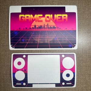 以下が含まれることがあります： レトロスタイルのステッカー2枚。上のステッカーには、夕焼けを背景にネオンフォントで「GAME OVER」と書かれています。下のステッカーは、ゲーム機に似た、ピンクと紫のアクセントが付いた白い中央部があります。
