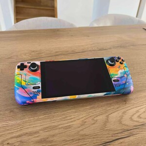 Op de afbeelding: Een draagbare gameconsole met een kleurrijk abstract ontwerp. De console heeft een zwart scherm, bedieningsknoppen en joysticks. De buitenkant is versierd met een levendige mix van blauw, geel, roze en groen, wat een unieke en artistieke uitstraling creëert.